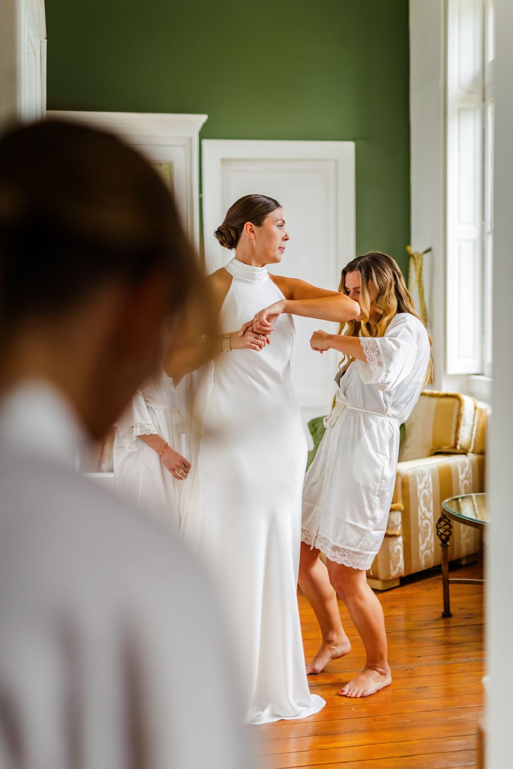 Two people in white robes prepare for a special occasion in a room with green walls and hardwood floors.