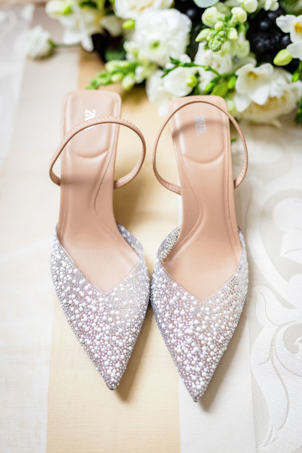 Sparkly silver rhinestone slingback heels with pointed toes displayed next to white flowers on a light background.