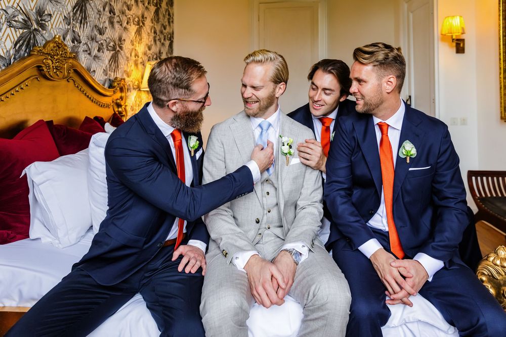 Groomsmen in navy suits and orange ties share a candid laughing moment on a couch at a wedding venue.