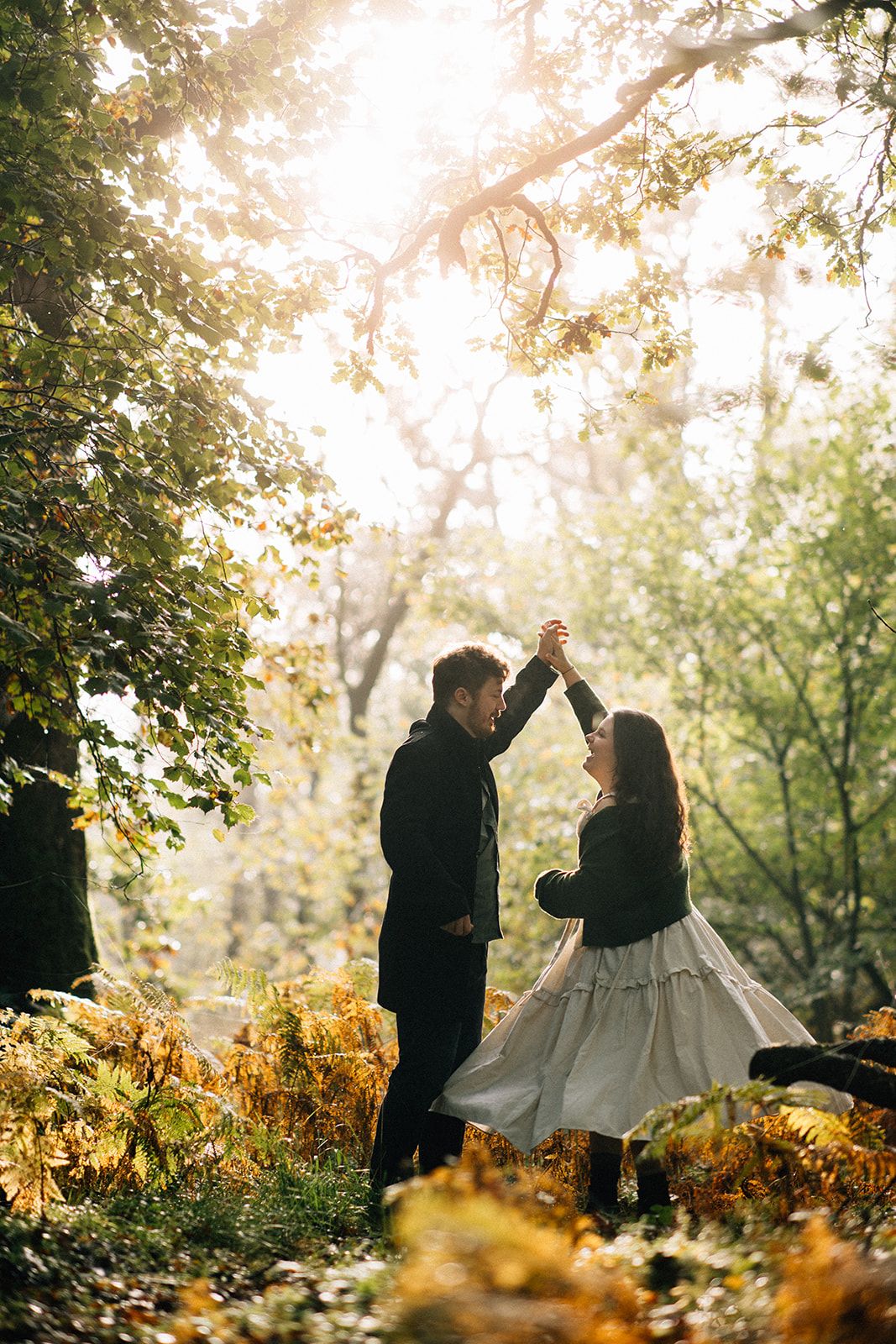 A couple dances together in a sunlit autumn forest setting with golden foliage and warm backlighting.