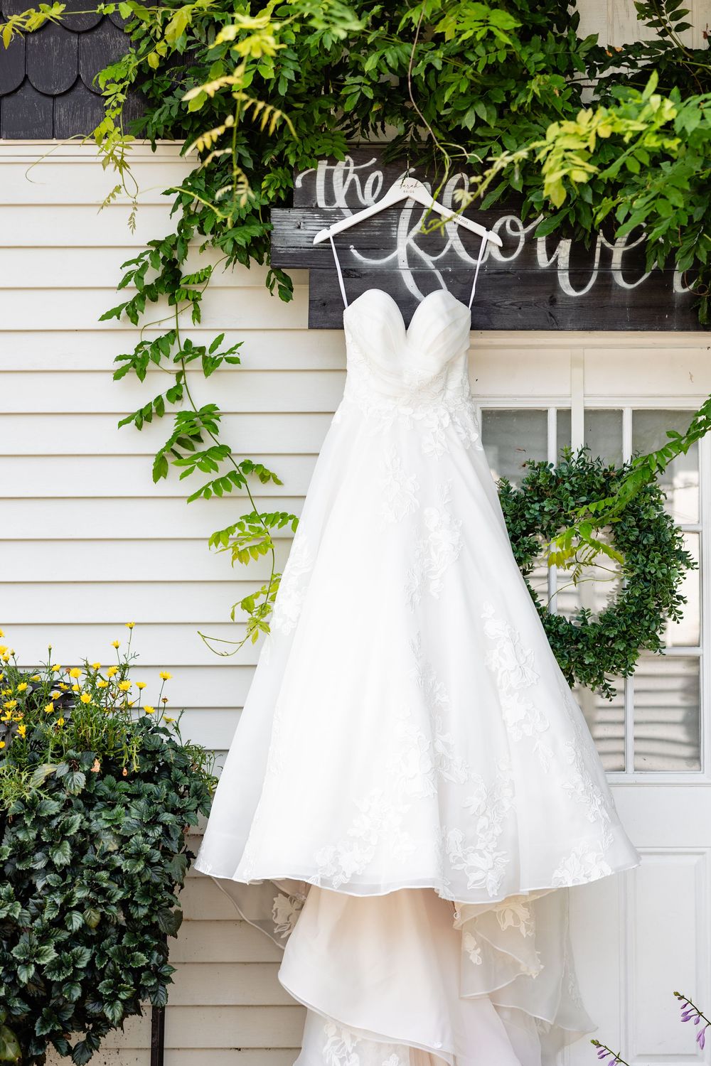 White lace A-line wedding dress hanging on custom hanger outside flower shop with green wreath and ivy backdrop.