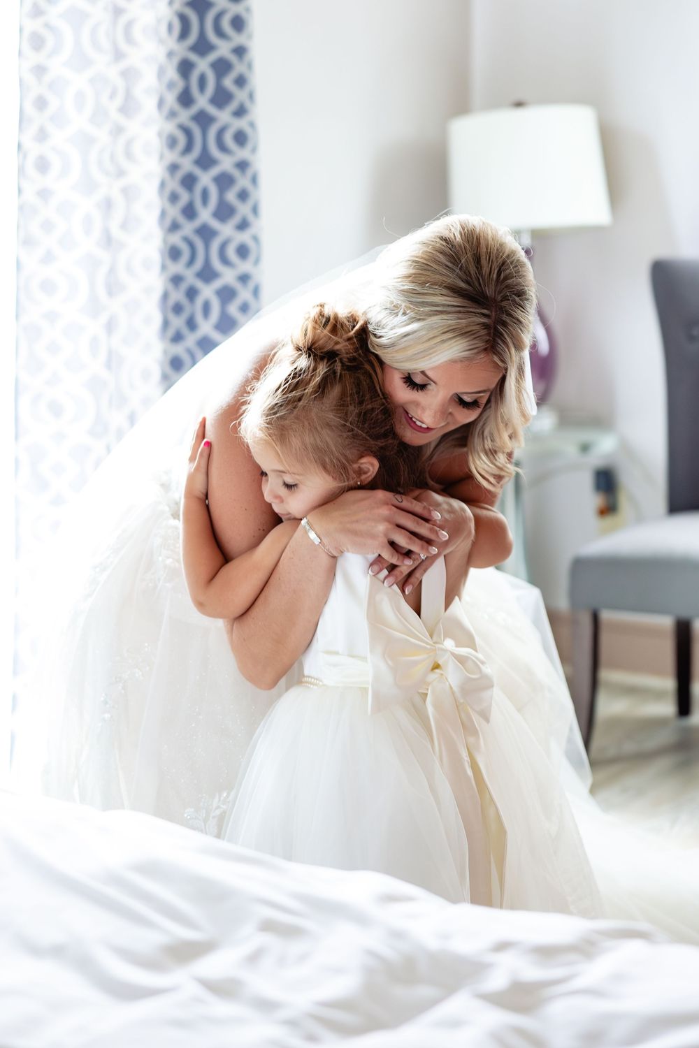 Bride in white wedding gown hugging young flower girl in matching white dress with bow, sharing tender moment in bright room.