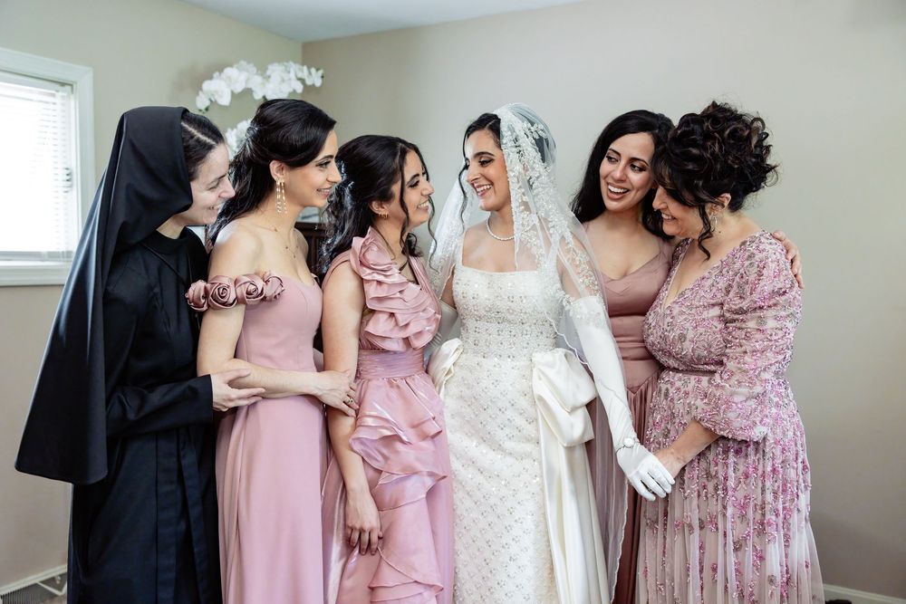Bride in white gown and veil smiles with bridesmaids in pink dresses and a nun habit in a bright room.