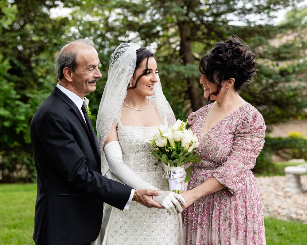 Bride in white gown and veil holds bouquet of roses while standing with two family members outdoors.