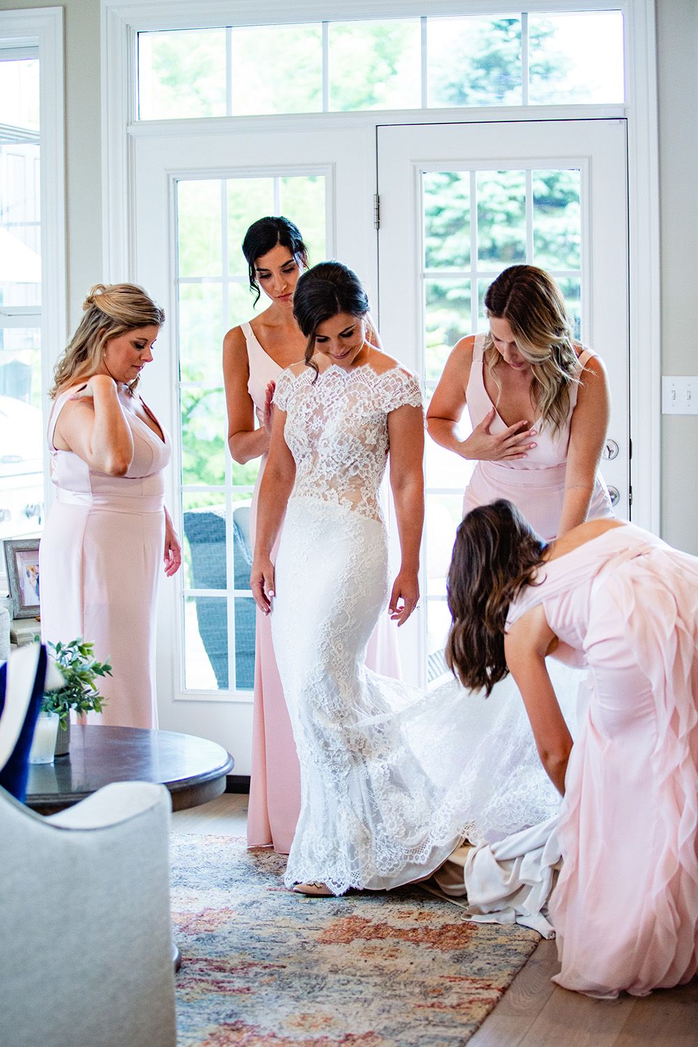 Bride in lace wedding gown surrounded by bridesmaids in blush pink dresses being helped to get ready indoors.