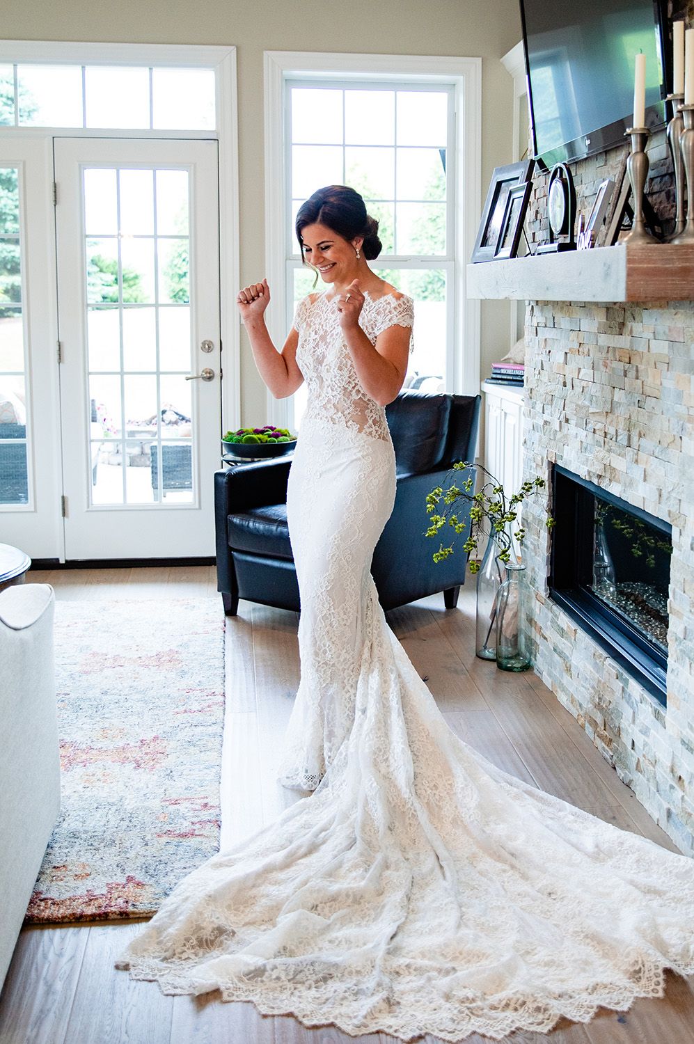 Bride in lace mermaid wedding gown with train poses excitedly in elegant home with stone fireplace.