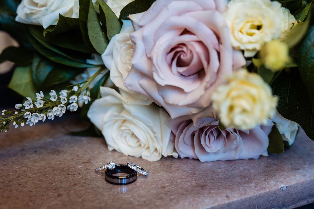 Wedding rings resting beside a bouquet of blush, cream, and lavender roses on a stone surface.