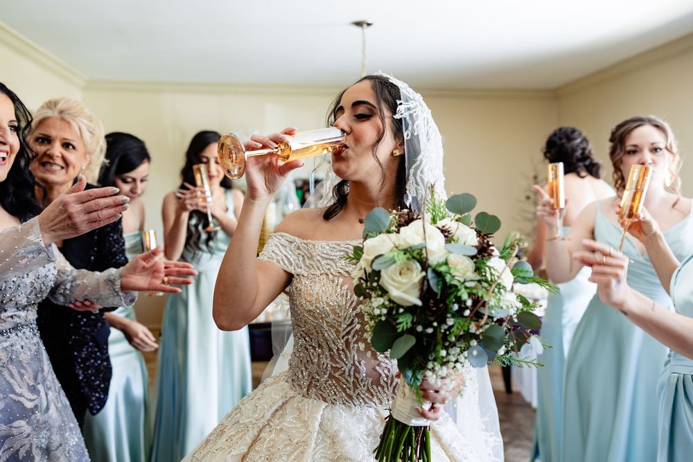 Bride in ornate gown drinks champagne with bridesmaids in blue dresses, holding white floral bouquet at wedding celebration.