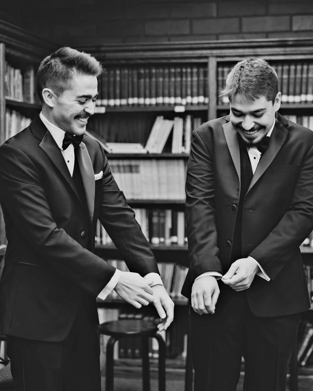 Two grooms in black tuxedos smiling while exchanging wedding rings in an elegant library setting.
