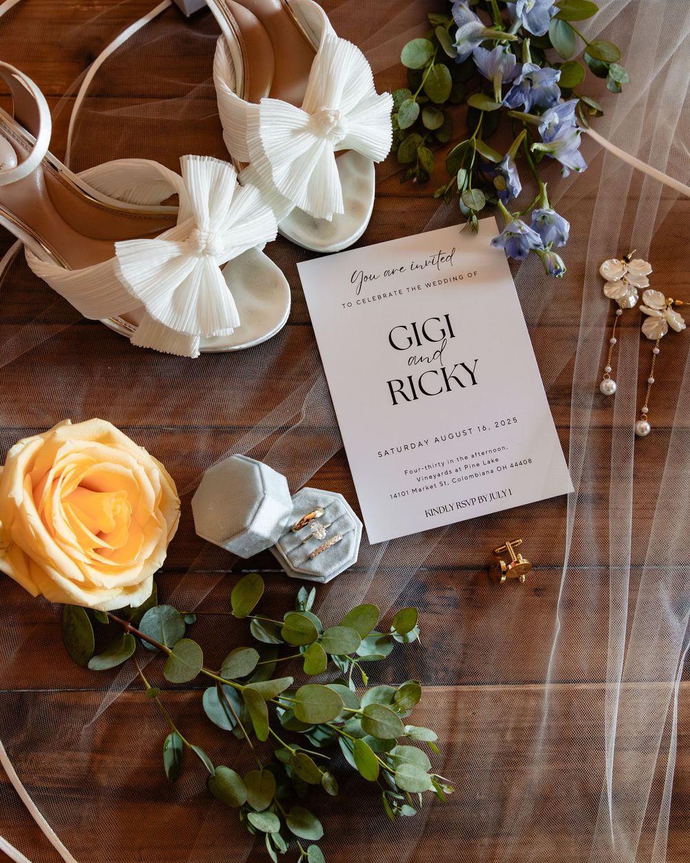 Wedding flat lay featuring Gigi and Ricky invitation, white bow heels, yellow rose, greenery, pearl earrings, and ring box on wood.