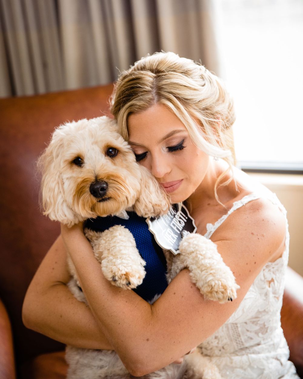 Bride in white lace dress tenderly holds a fluffy Cavapoo dog wearing a navy blue bow tie on her wedding day.