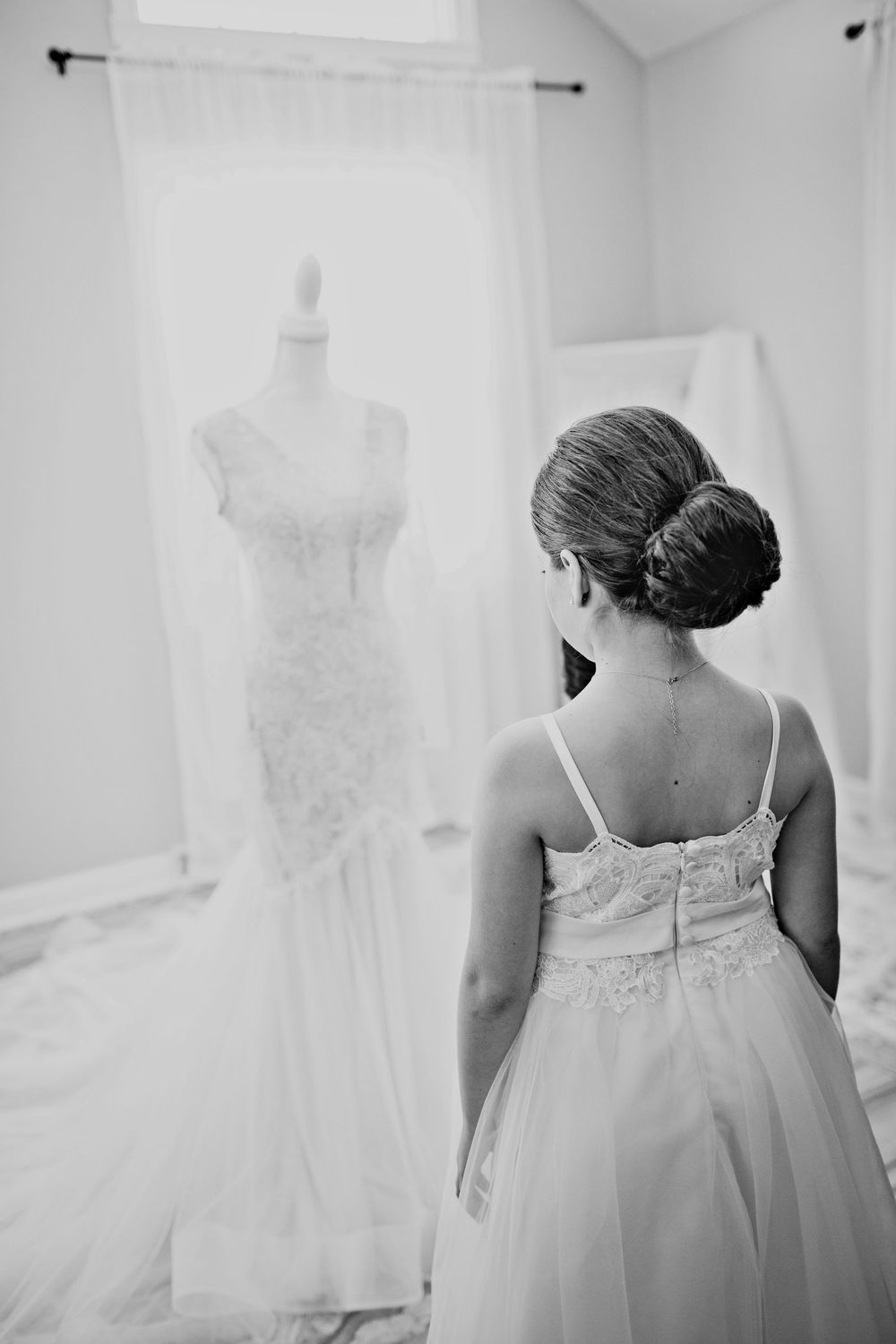 Bride in elegant gown views her wedding dress on a mannequin in a bright bridal suite, black and white photo.
