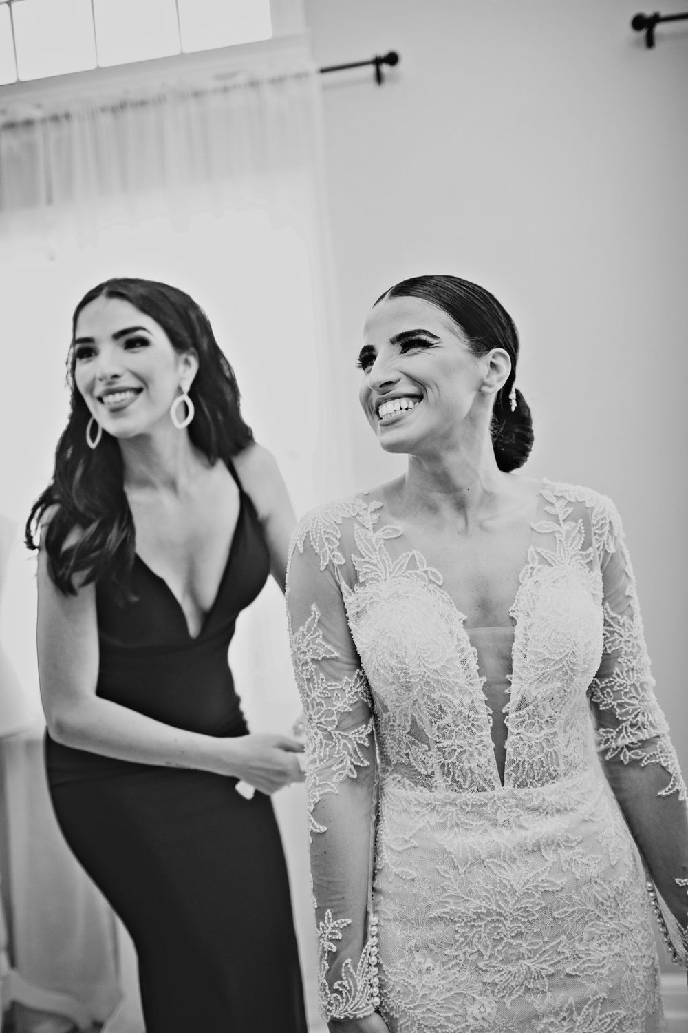 Black and white photo of a smiling bride in lace gown with bridesmaid in black dress at wedding.