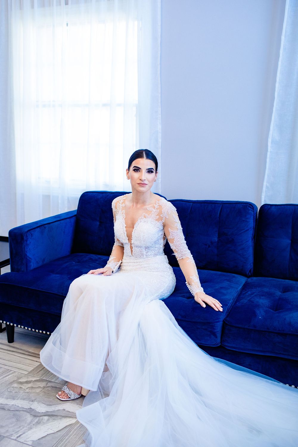 Bride in white lace long-sleeve gown sitting on blue velvet sofa near window in elegant bridal suite.