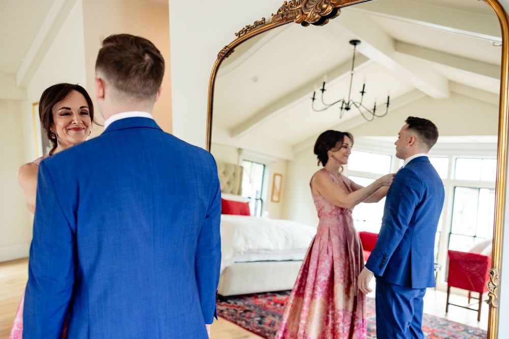 Couple in formal wear reflected in ornate gold mirror, man in blue suit helping woman in pink floral gown.