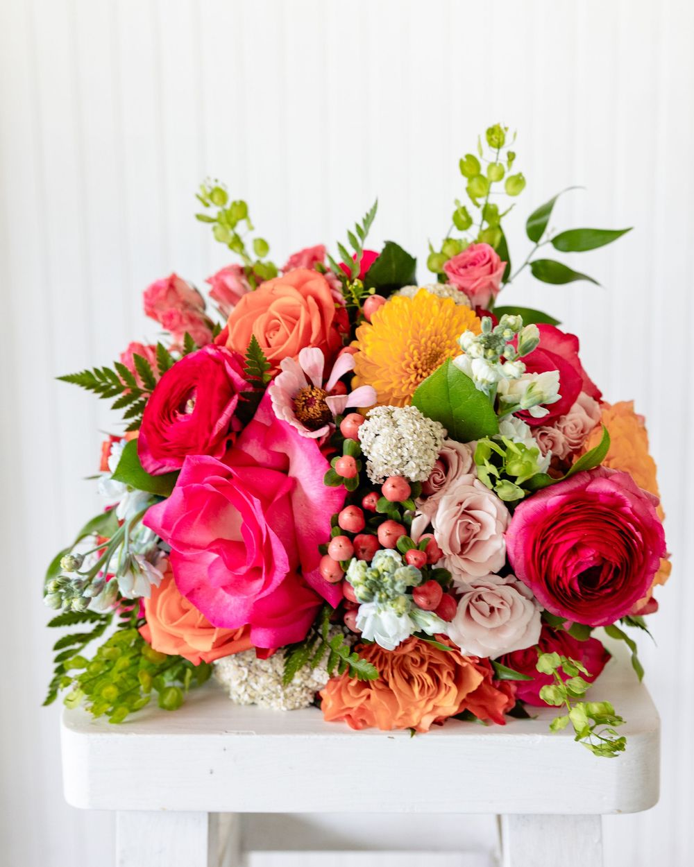 Vibrant mixed floral bouquet featuring pink roses, ranunculus, orange blooms, green berries, and lush foliage on a white stool.