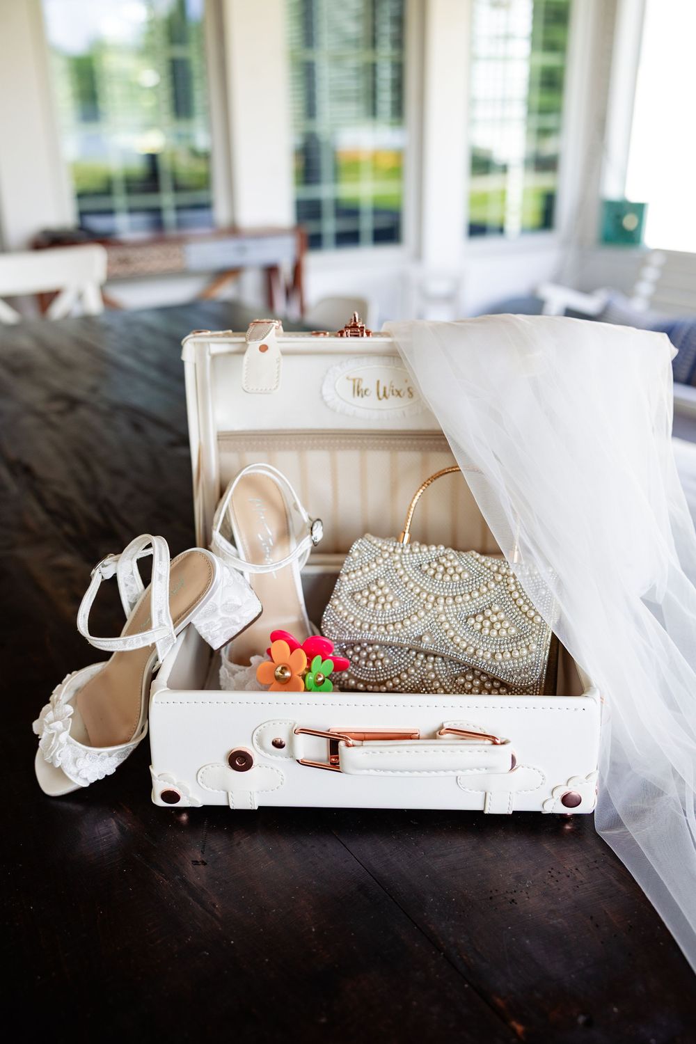 White vintage suitcase with bridal accessories including pearl clutch, white heels, and floral brooch on dark wood table.