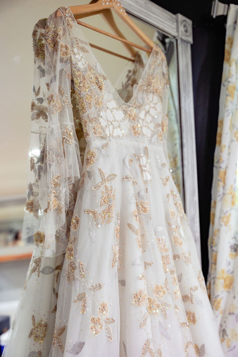 Elegant white wedding gown with gold floral embroidery and sheer long sleeves hanging on a wooden hanger.