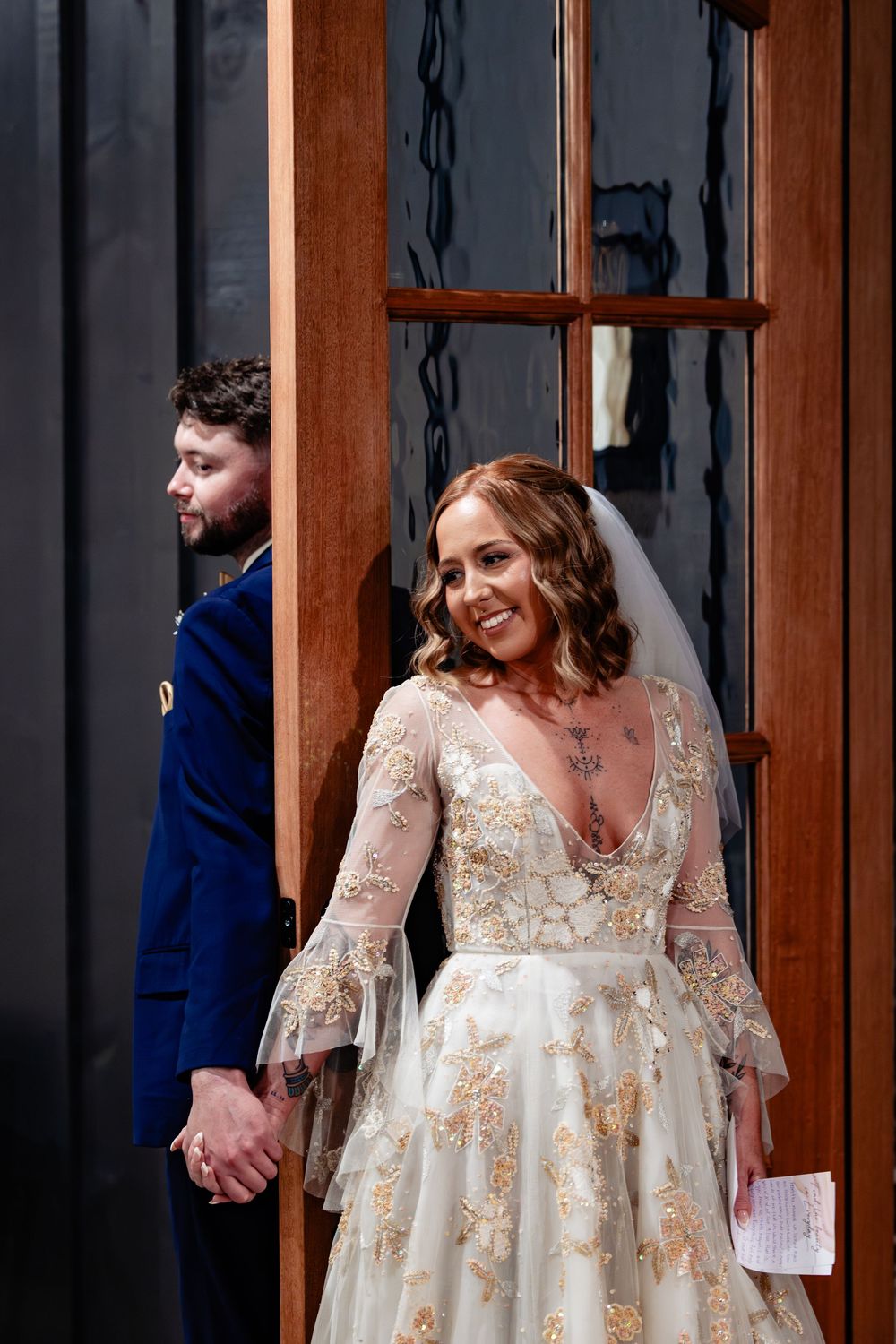Smiling bride in lace gown with veil holds hands with groom in blue velvet suit near wooden doors.