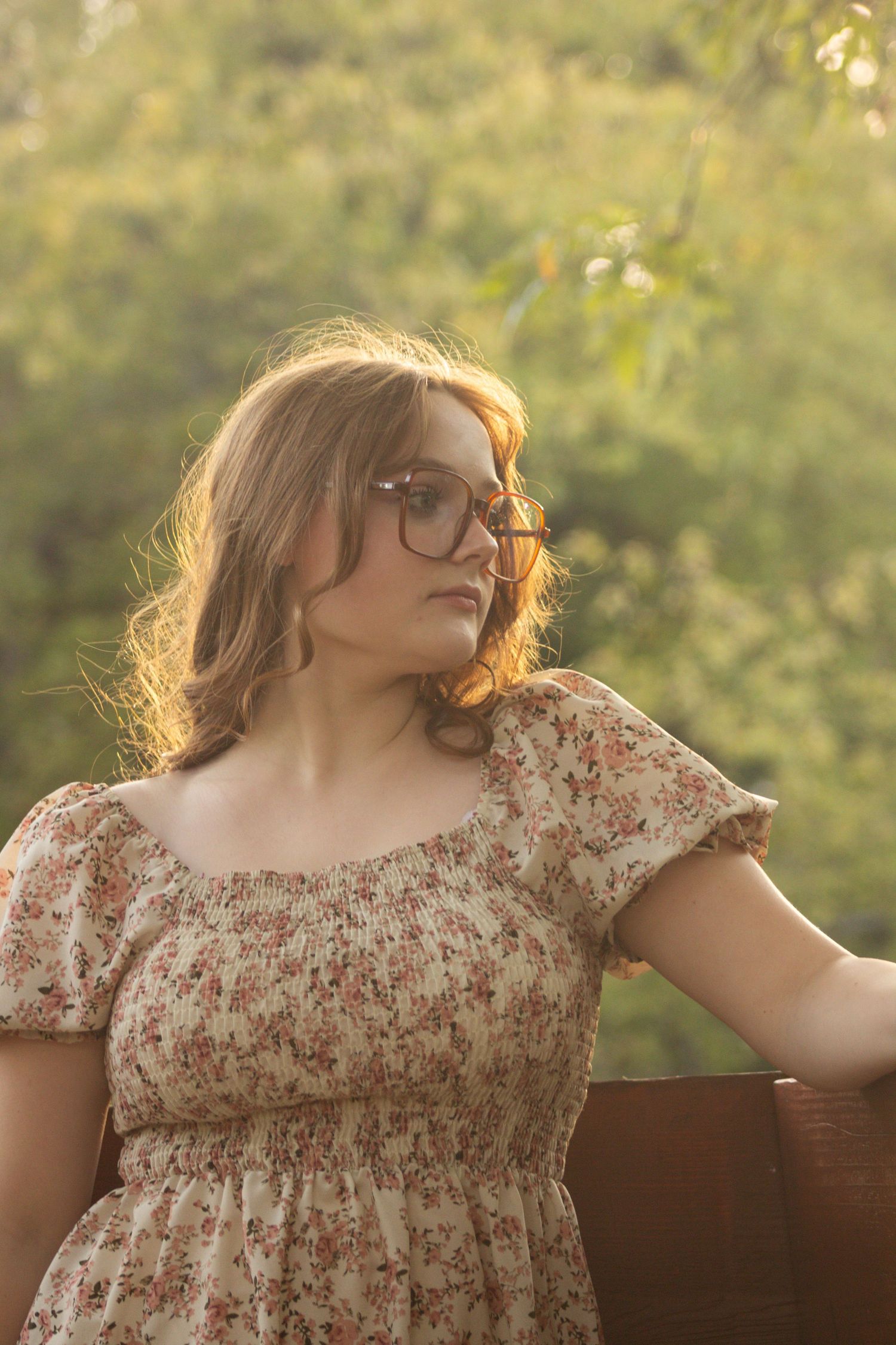 Person in a floral dress sits on a wooden bench during golden hour in a garden setting.