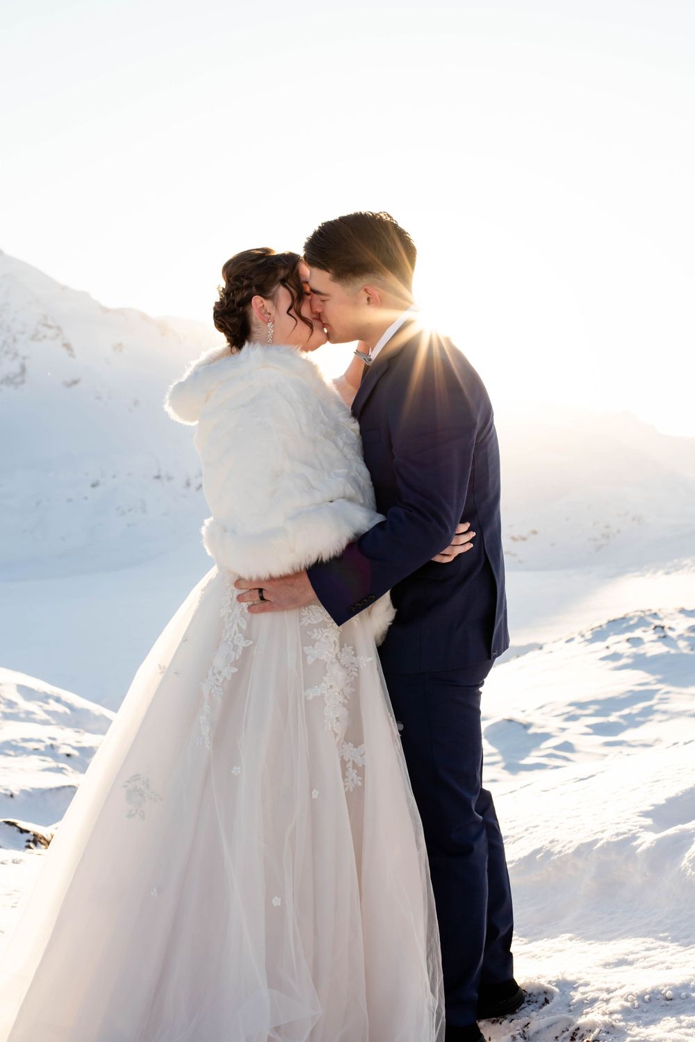 Winter wedding couple embracing in snow with sunburst behind them in mountains.