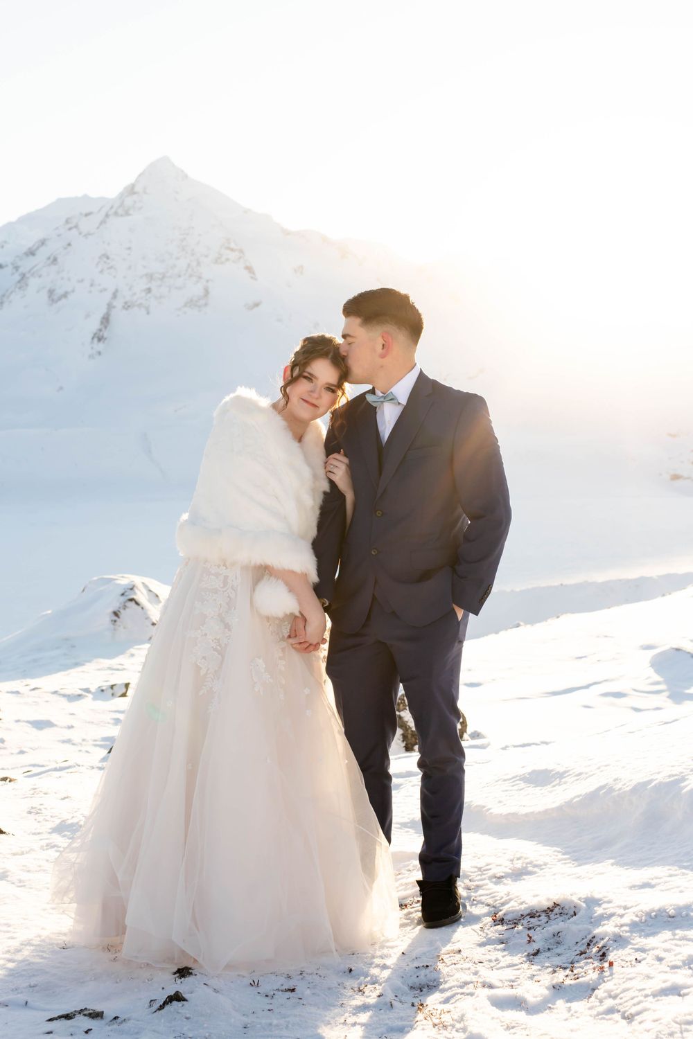 Romantic wedding photo of couple in formal attire against snowy mountain landscape.