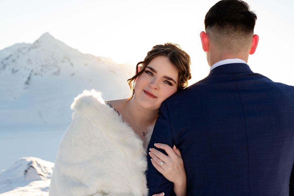 Bride in white fur wrap and groom pose against snowy mountain backdrop.