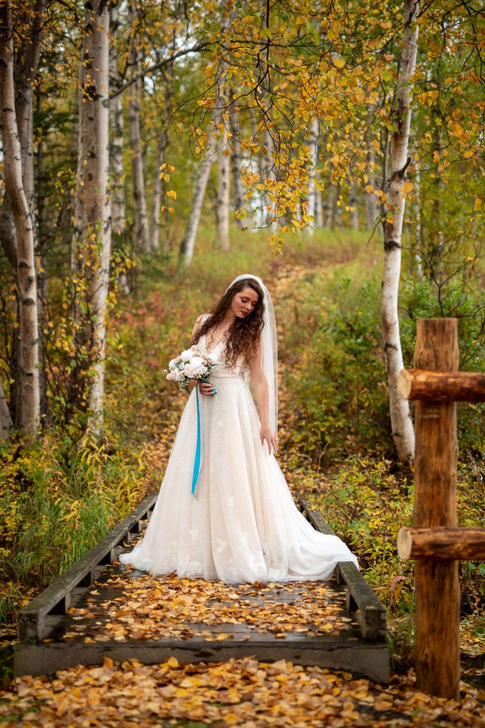 A bride in a white gown stands on a wooden walkway surrounded by fall foliage.
