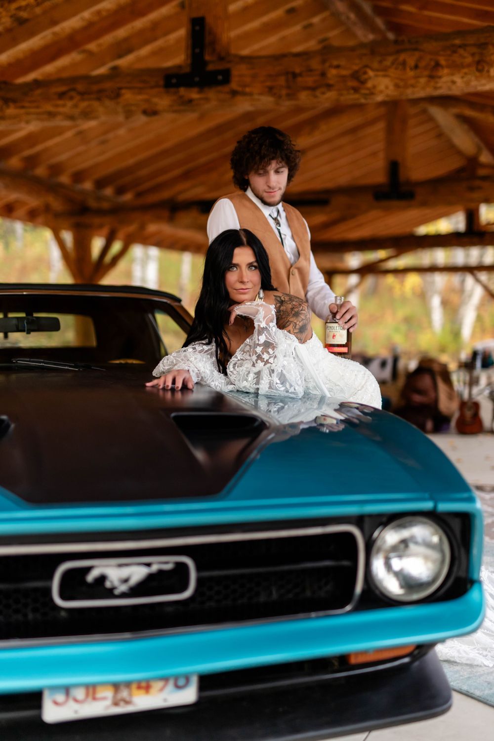 A stylish couple poses with a classic blue Ford Mustang under a wooden pavilion.