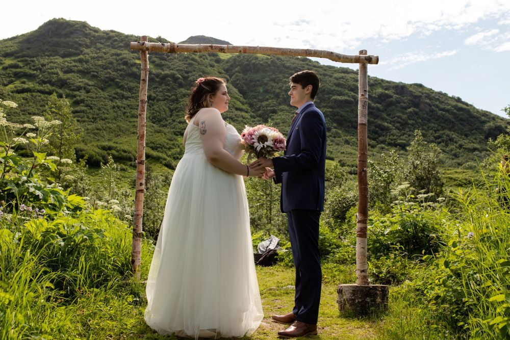 A wedding ceremony beneath a rustic wooden arch with mountains and greenery in the background.
