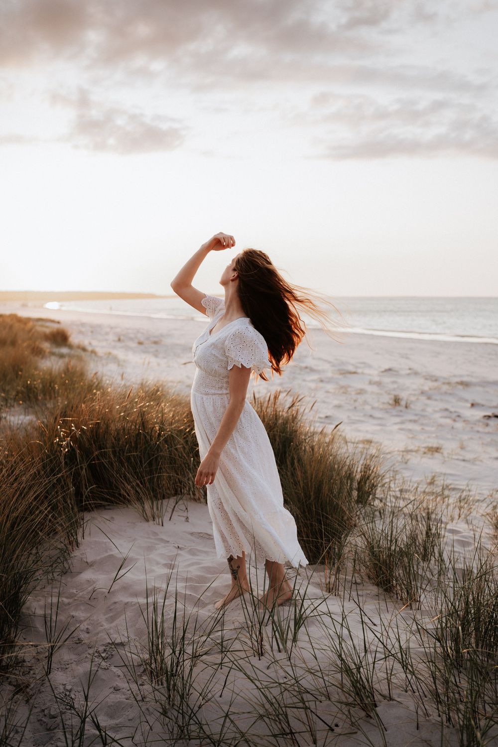 Person in white dress dancing on beach dunes at sunset with flowing movement.