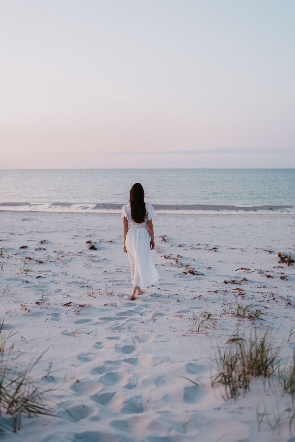 Figure in flowing white dress walks along beach at sunset leaving footprints in pristine sand.
