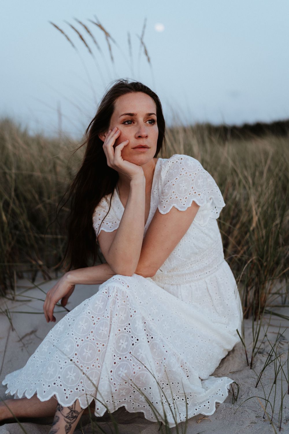 Figure in white eyelet dress poses among sand dunes at twilight.