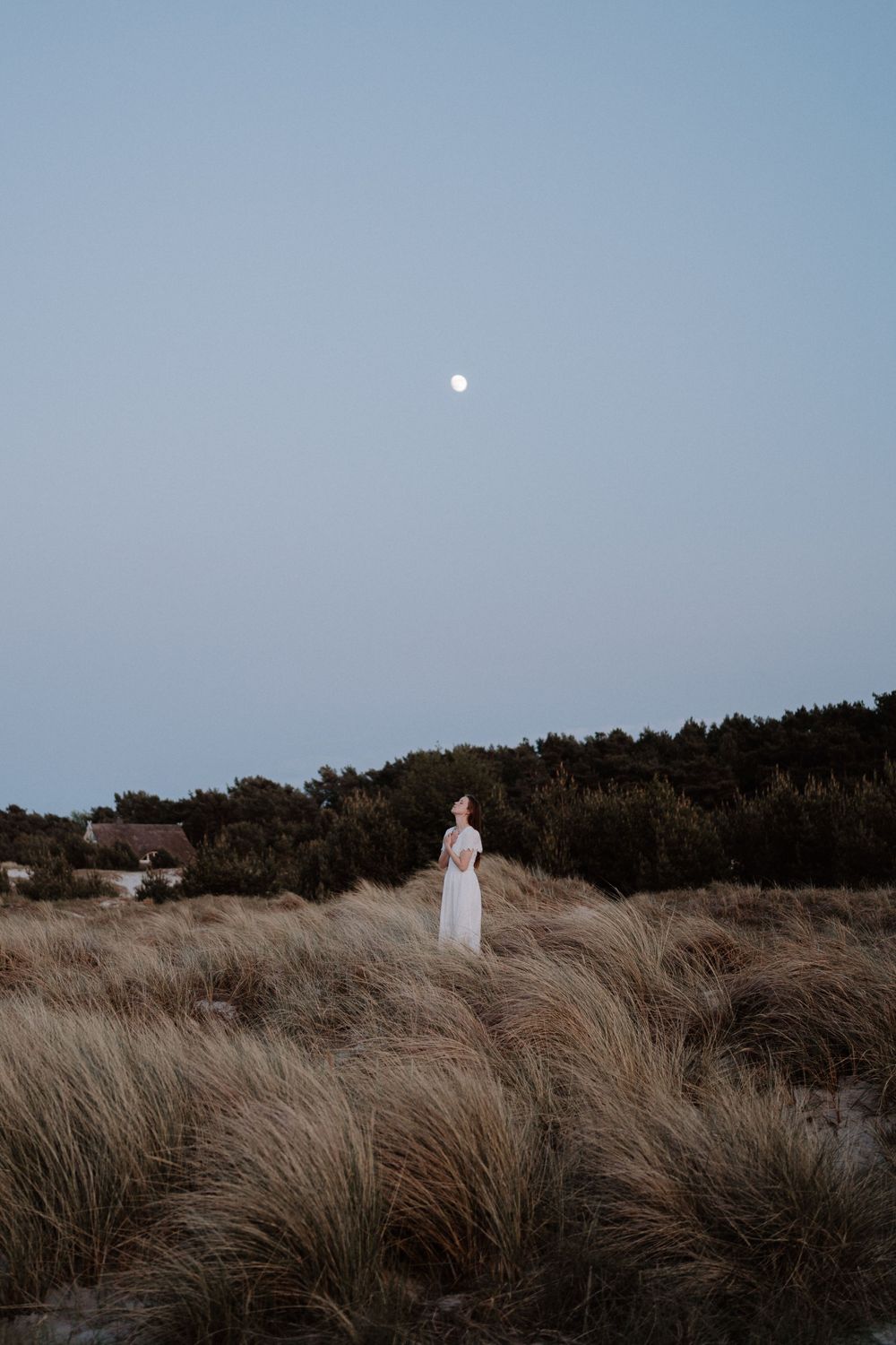 White dress contrasts against tall beach grass with moon glowing in dusky sky.