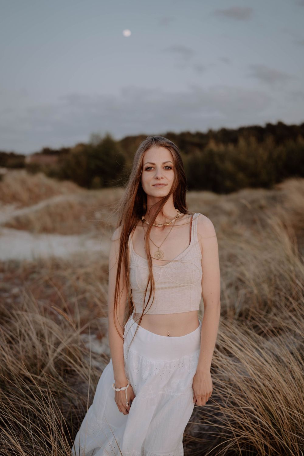 Figure in white outfit poses amid golden beach grass at dusk.