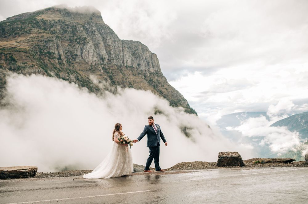 wedding couple walking along going to the sun road with moody clouds in the background, mountains playing peek-a-boo through the clouds