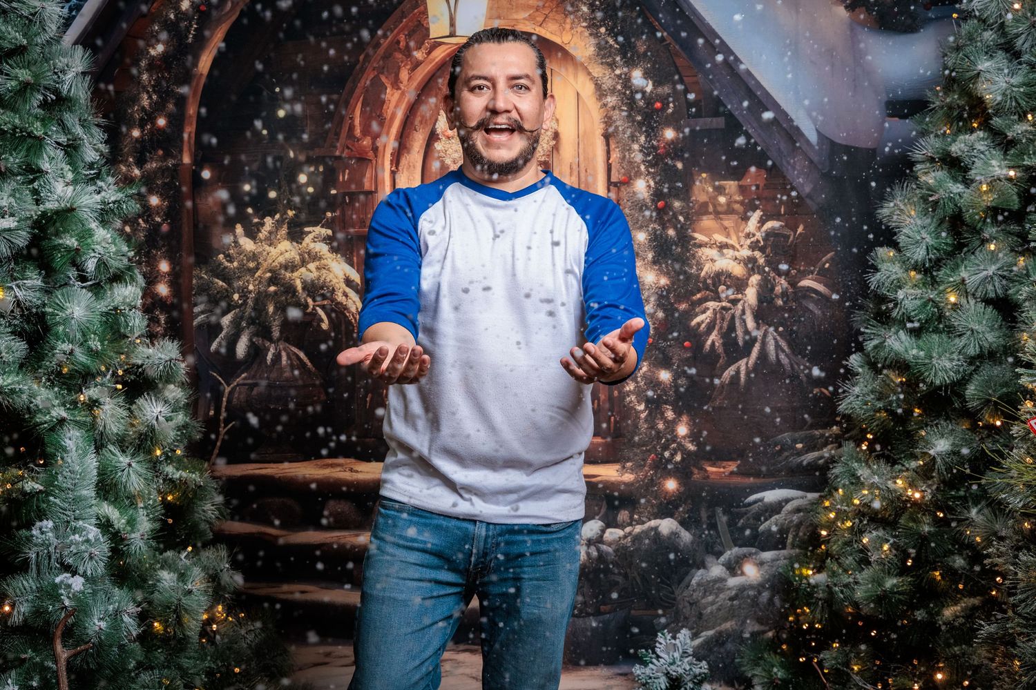 Person in a white t-shirt and jeans poses against a festive backdrop with Christmas trees and sparkling lights.