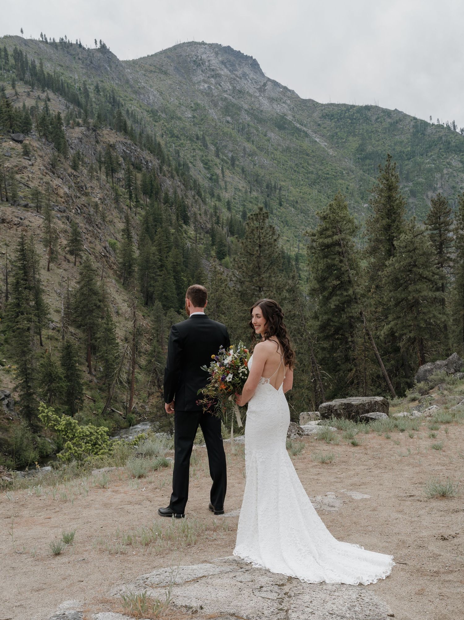 A couple shares a private moment in the mountains surrounded by evergreen trees and rugged terrain.
