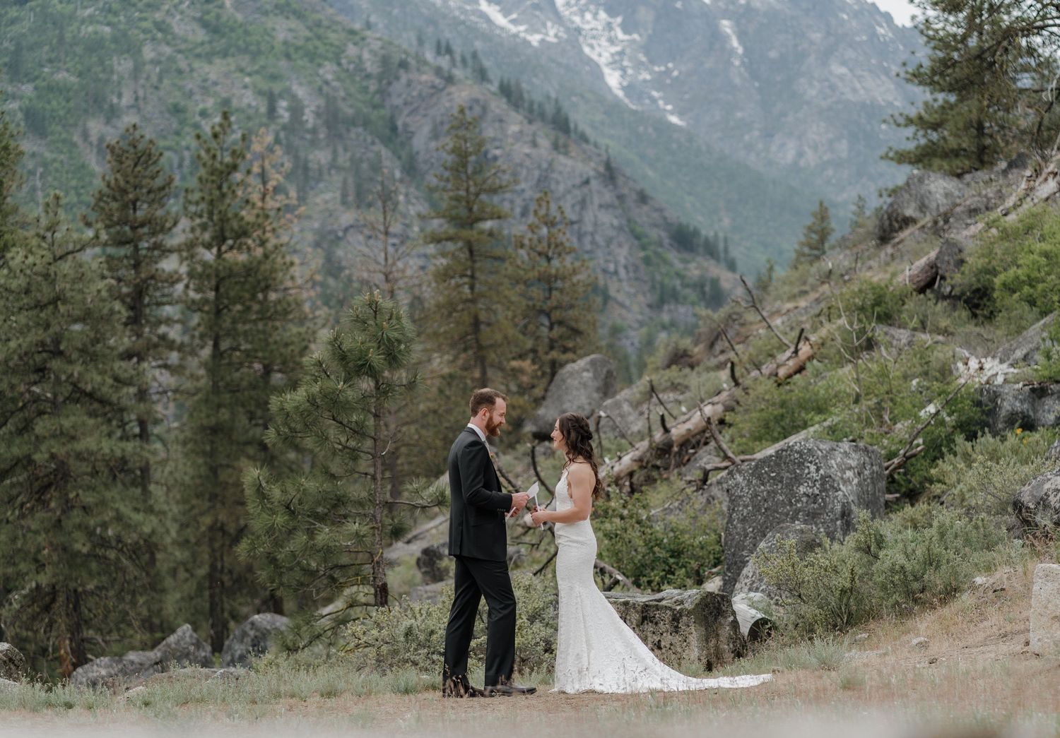 A couple in formal attire stands together in a scenic mountain landscape surrounded by evergreen trees and rocky slopes.