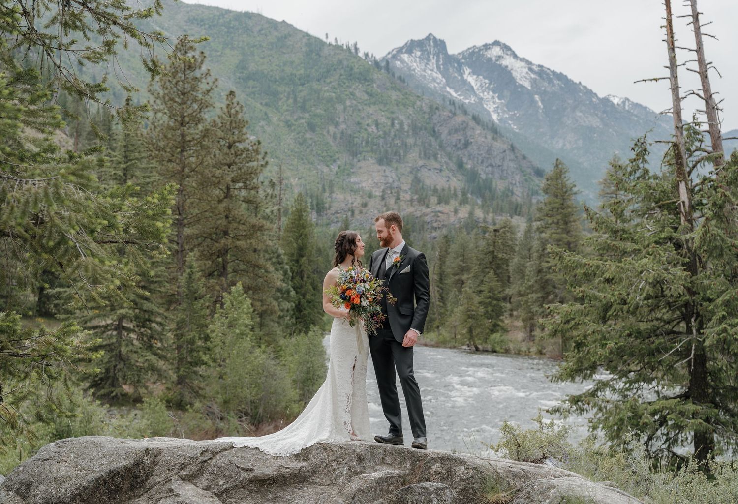 A couple in wedding attire stands among tall evergreen trees with majestic mountain peaks in the background.