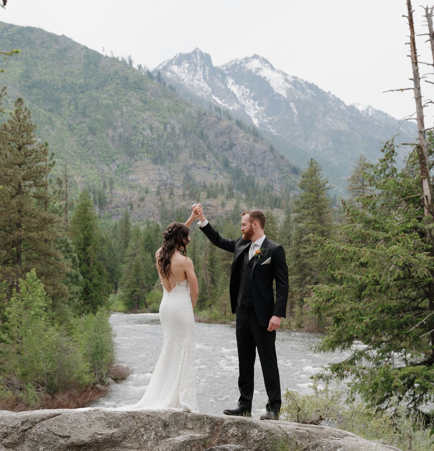 Couple in formal attire sharing an intimate moment against a backdrop of snow-capped mountains and evergreen forest.