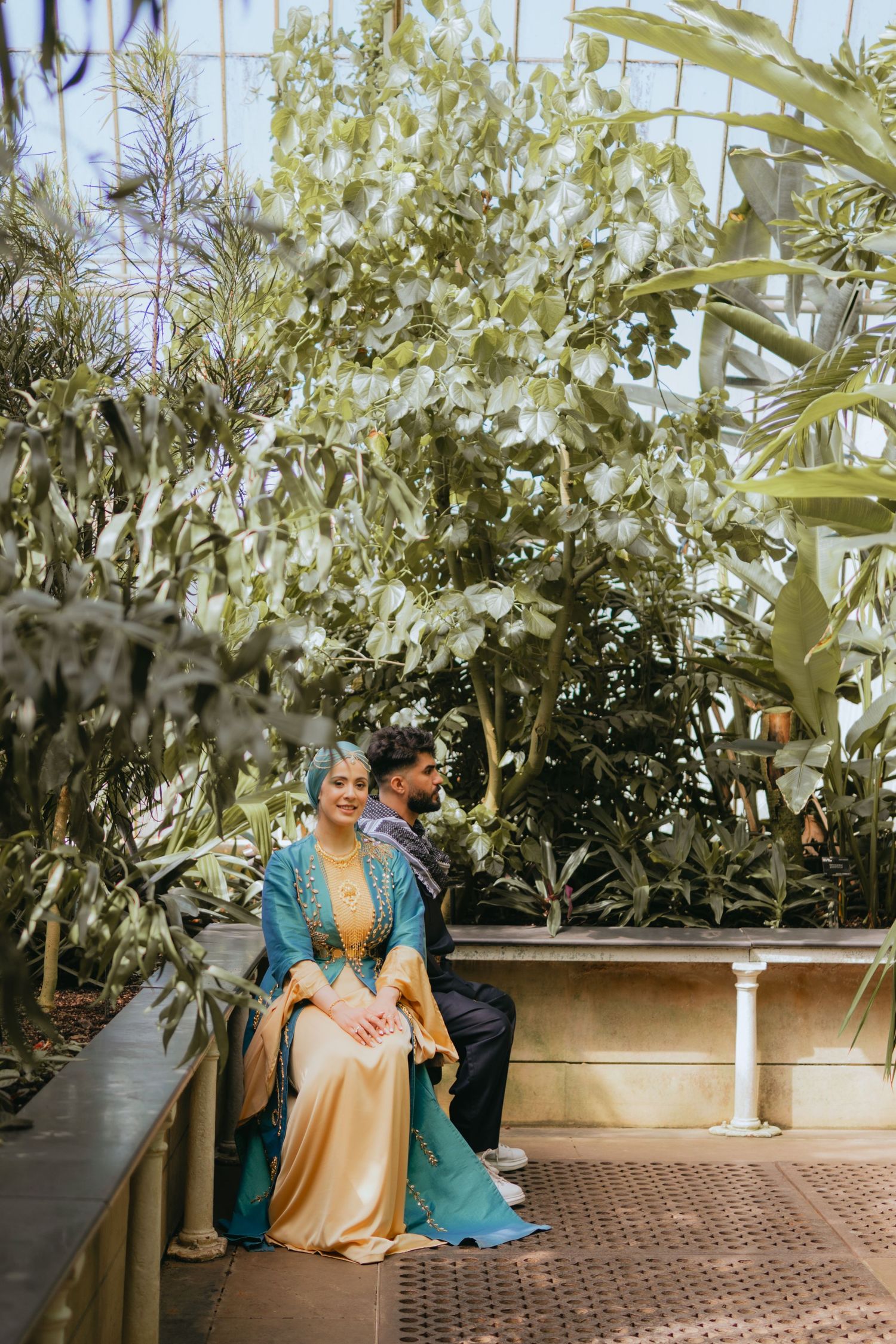 A couple stands together on a wooden boardwalk surrounded by lush greenery and willow trees in a garden setting.