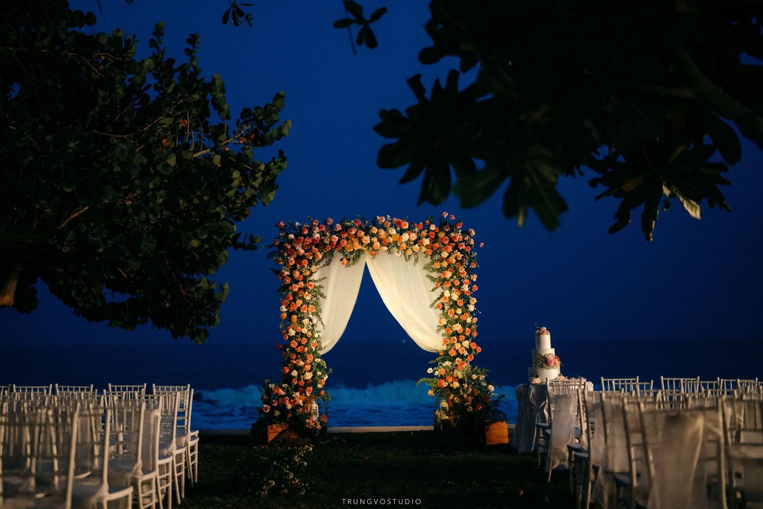 Elegant outdoor wedding ceremony setup with floral arch and white chairs at night by the sea.
