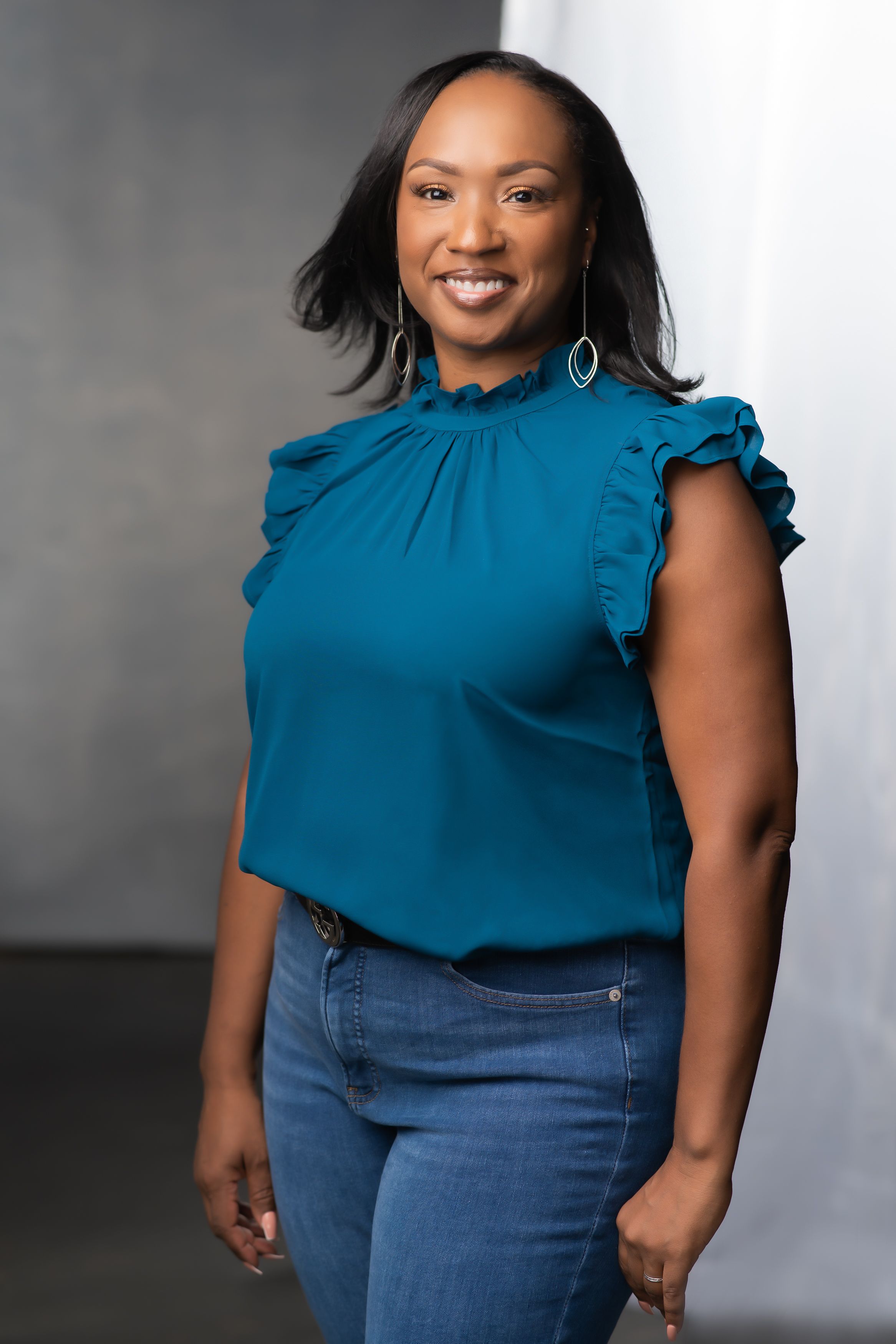 A model in a teal ruffled blouse and blue jeans poses confidently against a gray studio backdrop.