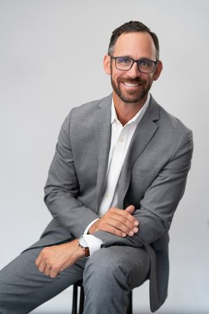 Professional headshot of person in gray suit and glasses sitting against light background.