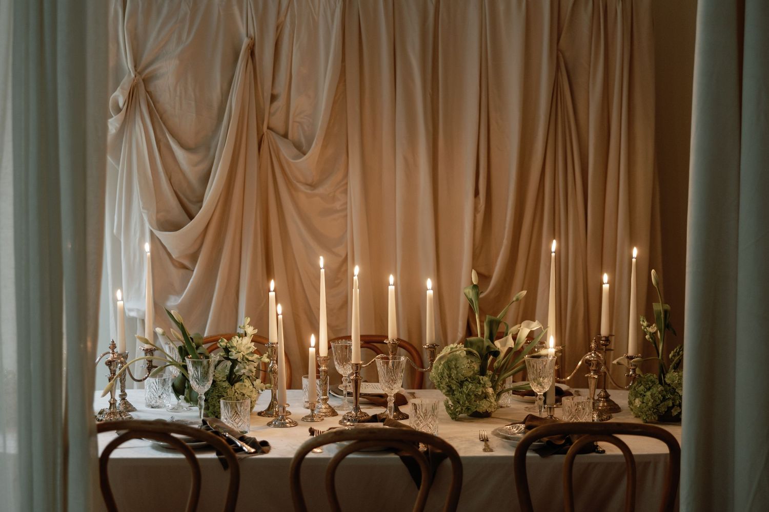 Romantic candlelit wedding reception table with bentwood chairs and flowing fabric backdrop.