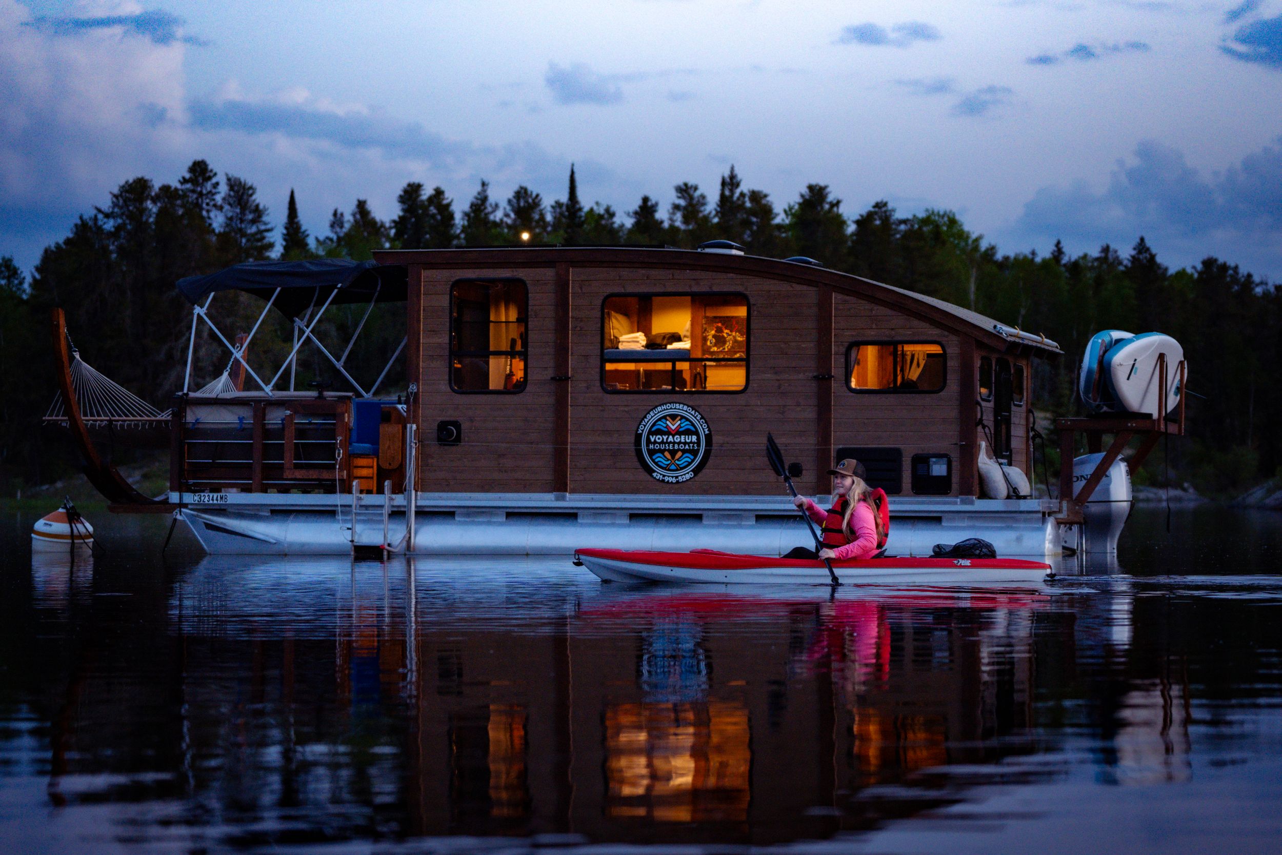 Illuminated houseboat on calm lake waters at dusk with kayaker paddling nearby.