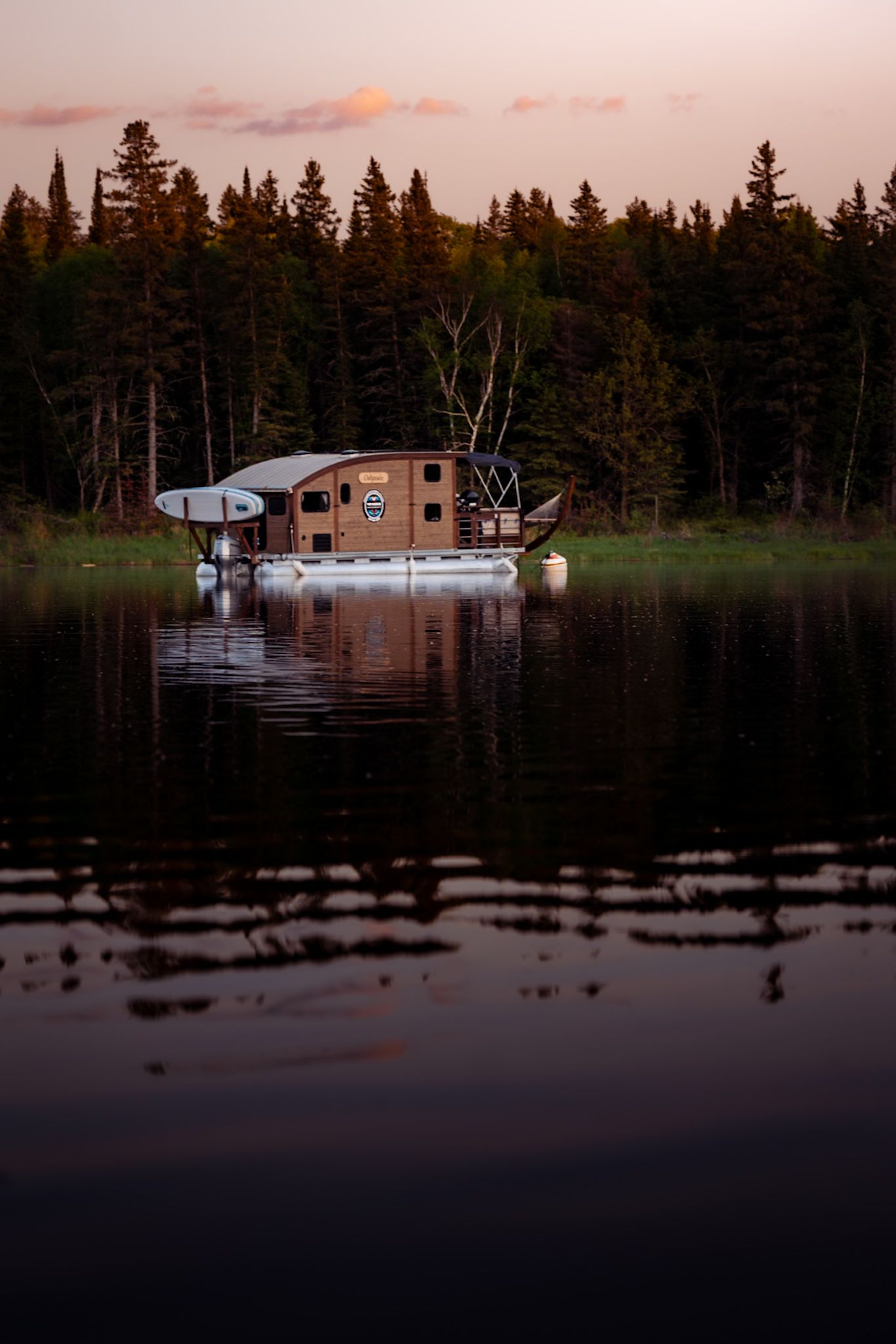 Houseboat moored on calm lake at sunset with pine forest backdrop.