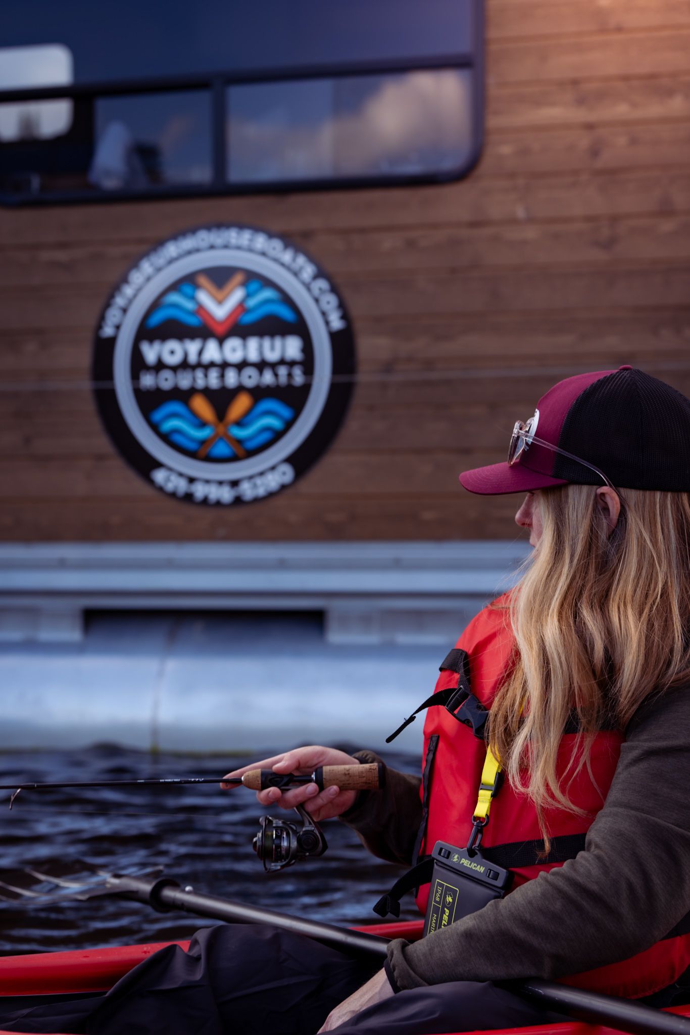 Angler in red life vest fishing from kayak near wooden boat with Voyageur Houseboats logo.