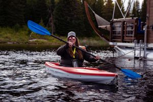 Kayaker paddling on calm lake waters near forest and dock during overcast day.